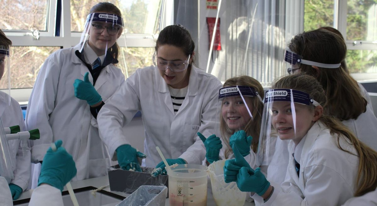 Five people in white lab coats, wearing face shields  and teal gloves , mixing chemicals in laboratory setting